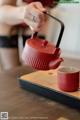 A woman pouring tea into a red teapot on a wooden tray.