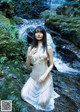 A woman in a white dress sitting on a rock by a waterfall.