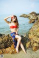 A woman in a red bikini sitting on a rock by the water.